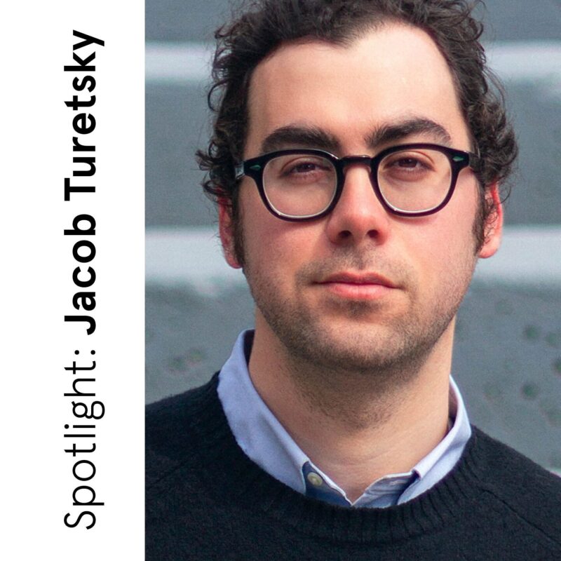 Portrait of Jacob Turetsky wearing round glasses and a dark sweater, standing against a muted gray architectural background, with “Spotlight: Jacob Turetsky” text along the side.