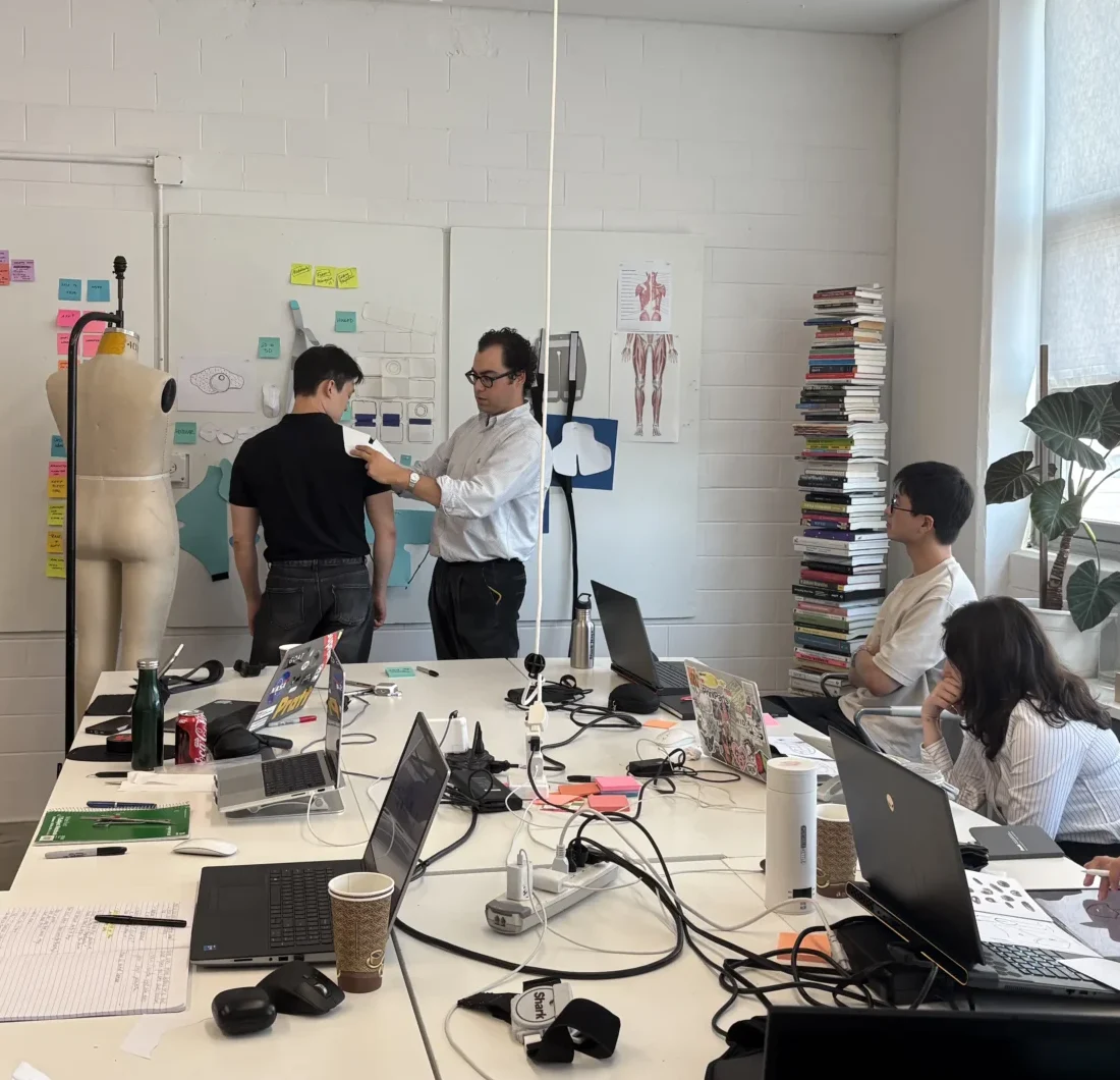 Interwoven Design Group team members reviewing a wearable prototype during a studio meeting, with anatomical references and design notes on the wall