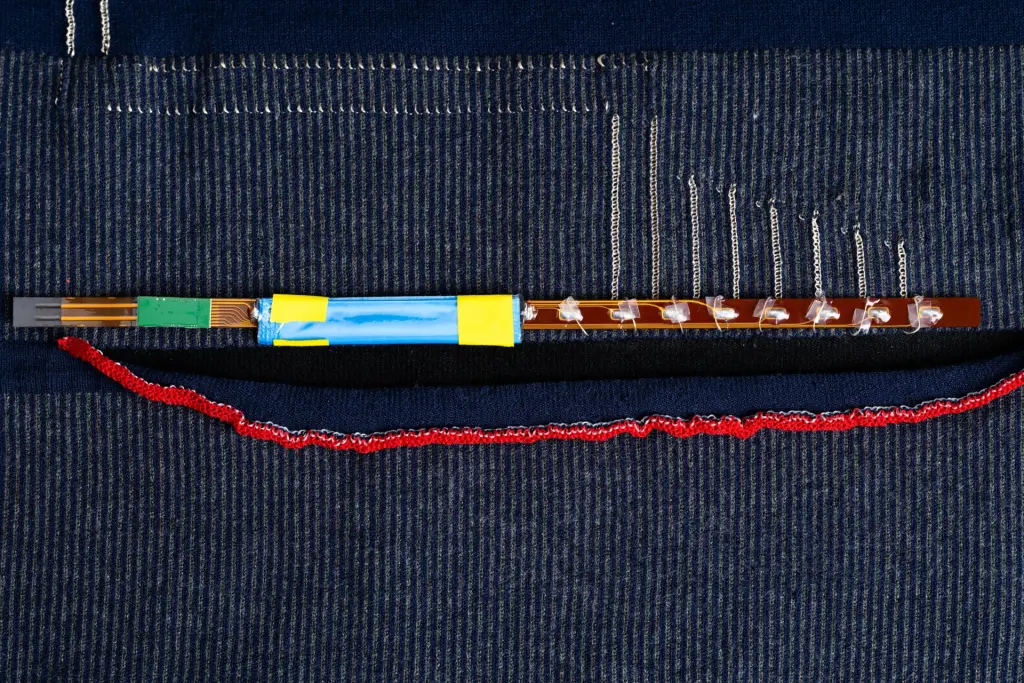 A flexible circuit with sensor pad, PCB, power module, and surface-mount components integrated into a knit textile alongside conductive threads and stitching, illustrating smart textile construction