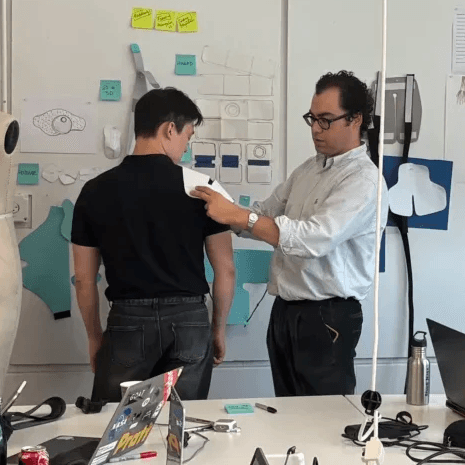 Designer adjusting wearable prototype on colleague in studio with whiteboard and laptops.